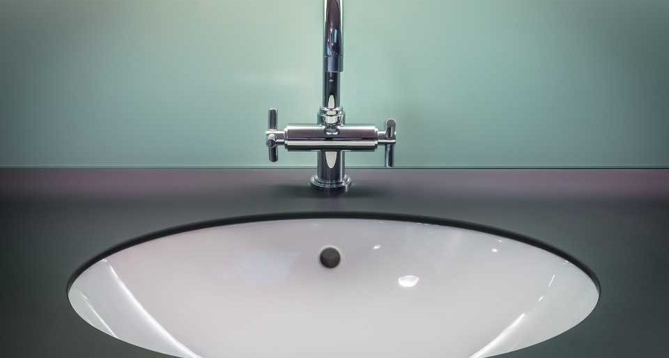 Weißes Keramikwaschbecken eingelassen in einen schwarzen Waschtisch mit einer silbernen Armatur vor blassgrüner Wand